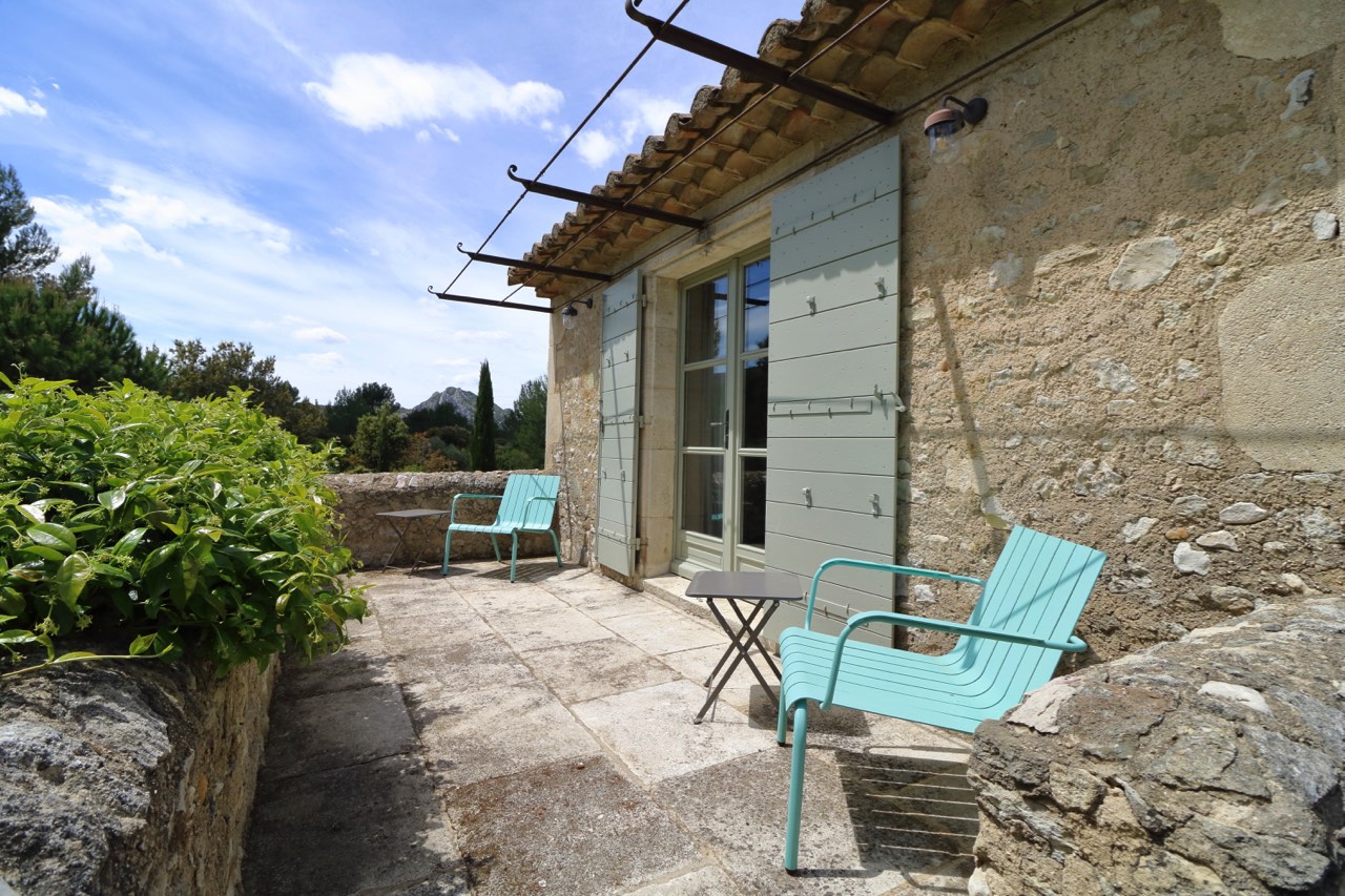 A serene patio area with two teal chairs and a matching table, nestled against an old stone house with green shutters, surrounded by lush greenery under a clear sky.